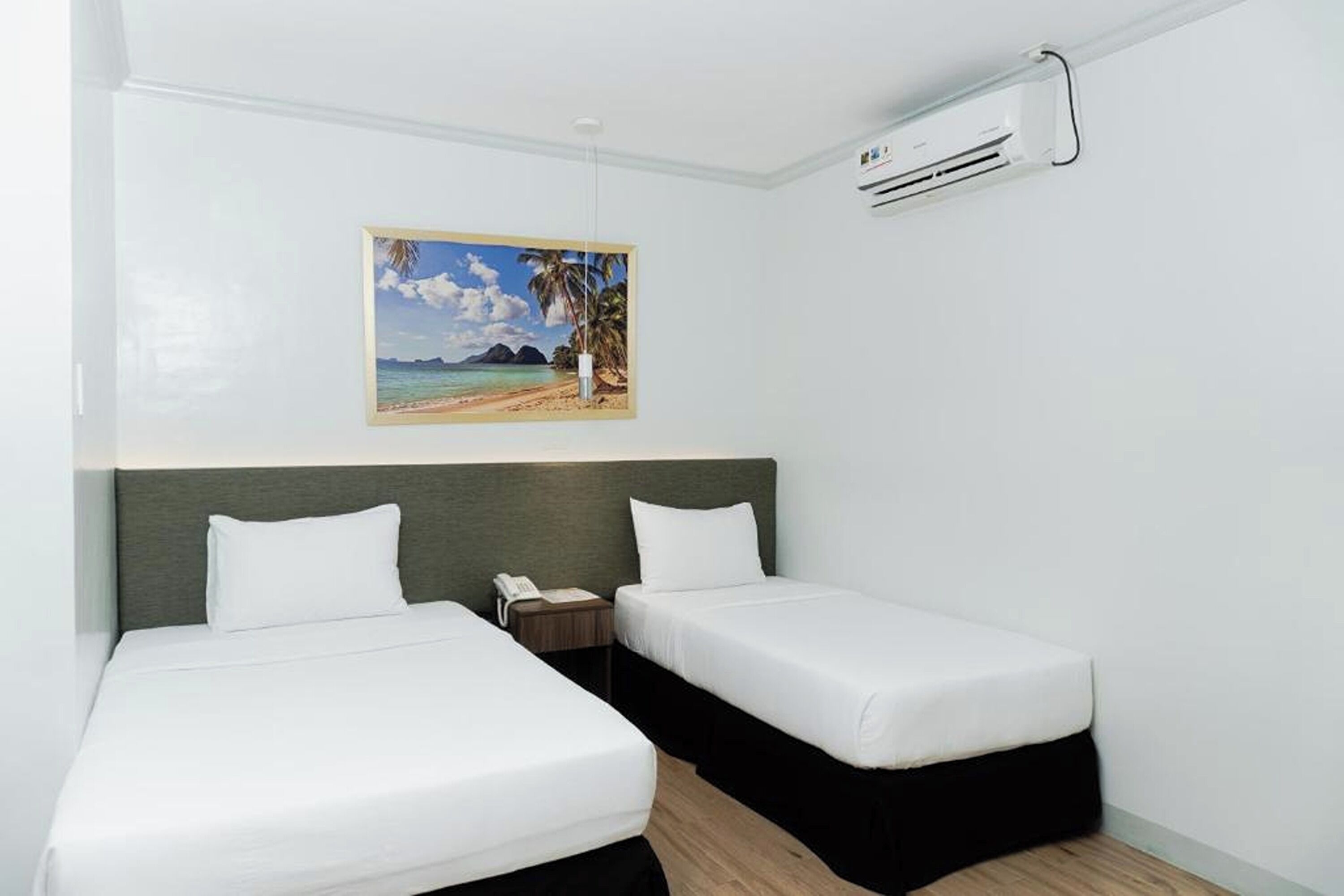 A hotel room with two beds, a painting of a beach, and an air conditioning unit.