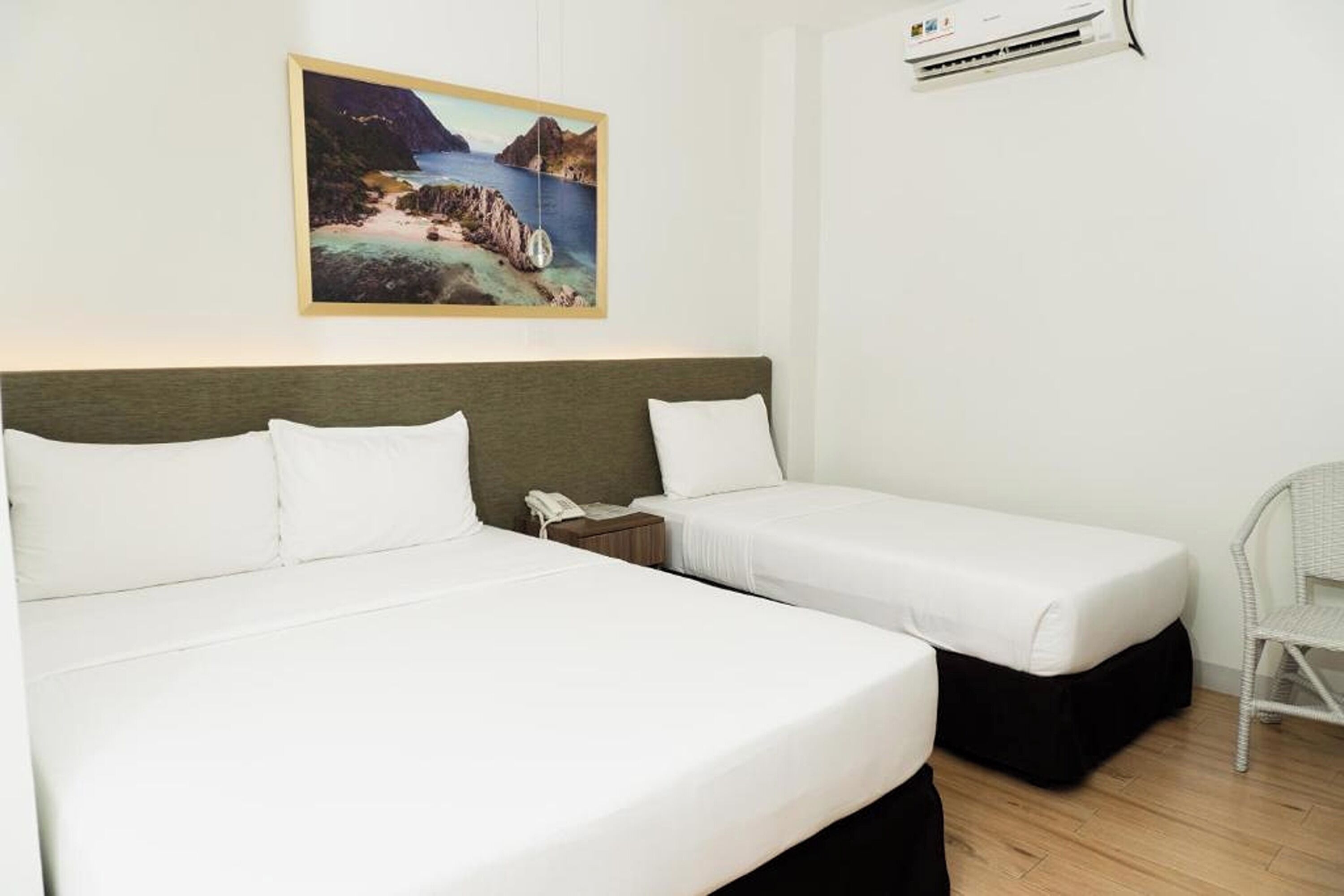 Habitación de hotel con dos camas, un cuadro de un paisaje costero, un equipo de aire acondicionado y una silla de mimbre.