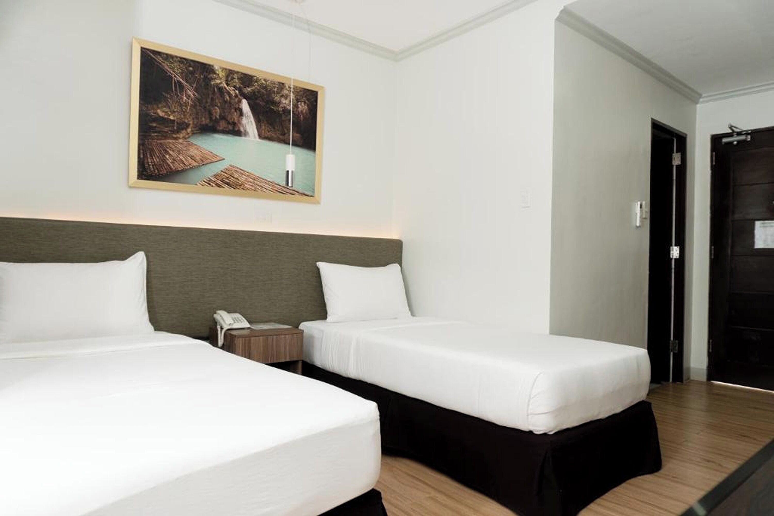 A hotel room with two beds, a painting of a waterfall, a bedside table with a phone, and a wooden floor.