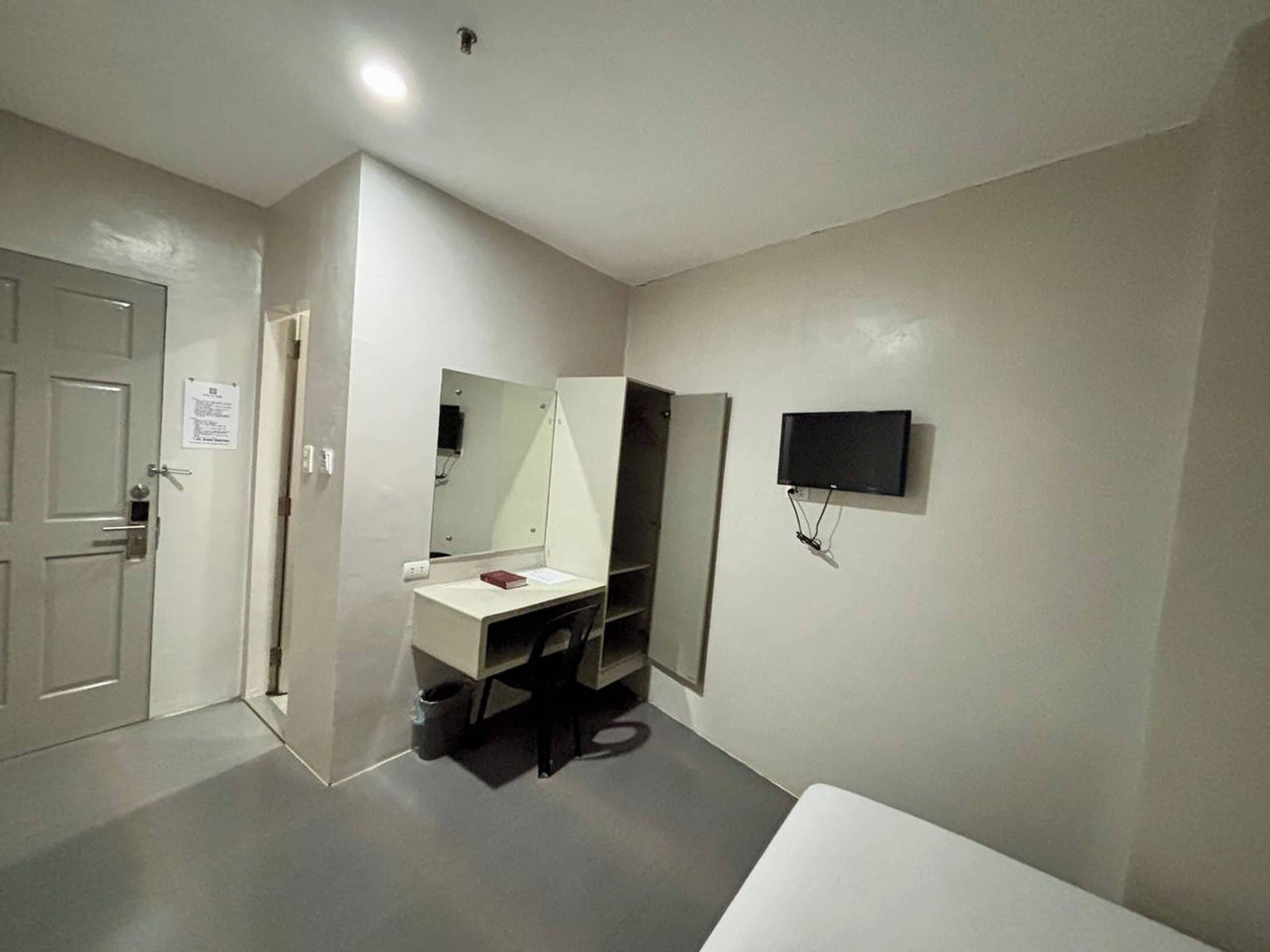 A small, modern bathroom with a white sink, a mirror, and a toilet. There is a door to the left and a television mounted on the wall.