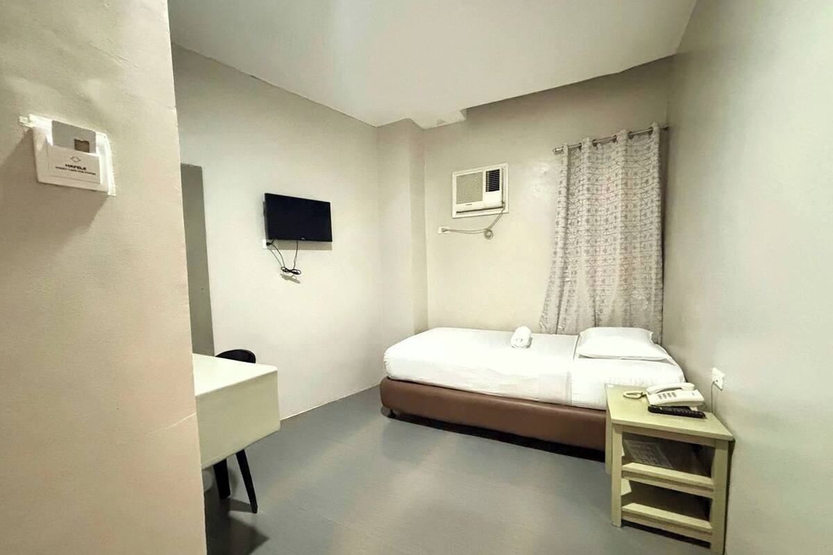 Ein kleiner, schlichter Raum mit einem Bett, einem Nachttisch, einem an der Wand befestigten Fernseher und einem Fenster mit Vorhang.