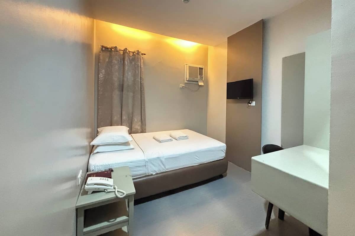 Ein kleines, schlichtes Hotelzimmer mit einem Einzelbett, einem Nachttisch mit Telefon, einem Schreibtisch und einem Stuhl.