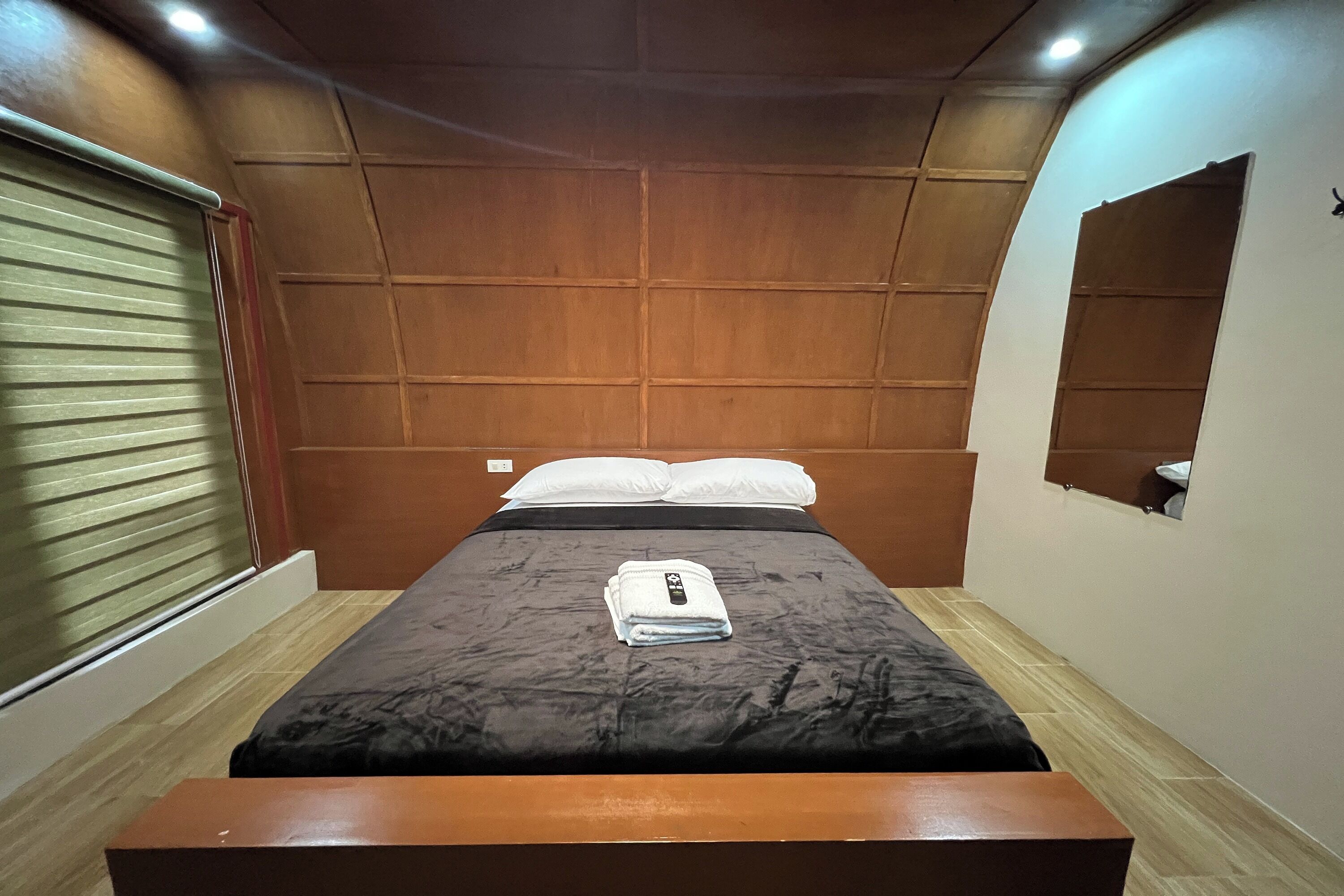 A bedroom with a bed, a bedside table, a window with blinds, and a mirror on the wall.