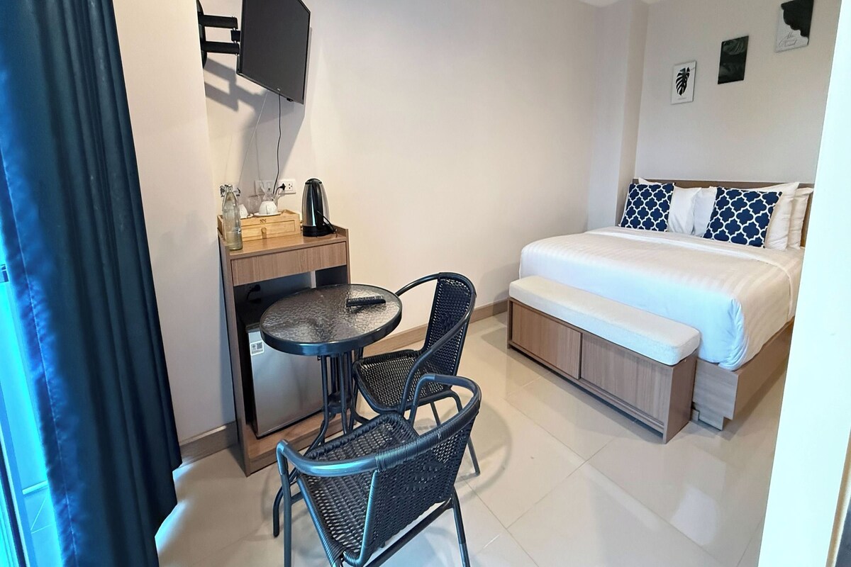 A compact hotel room with a bed, a desk with a chair, a small table, a TV mounted on the wall, and a few wall decorations.
