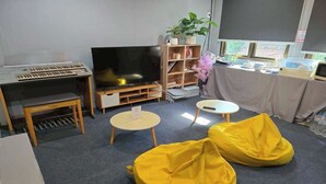 A room with a piano, a large flat-screen TV, two yellow bean bags, a small table, and a bookshelf with plants.