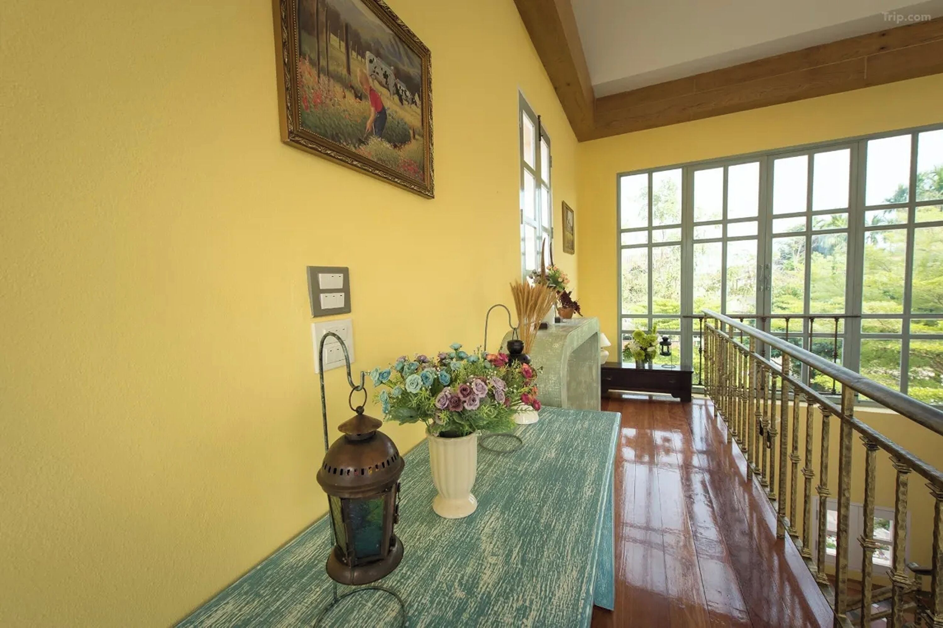 Una habitación con pared amarilla, un ventanal grande, barandilla de madera y una mesa con un jarrón de flores.