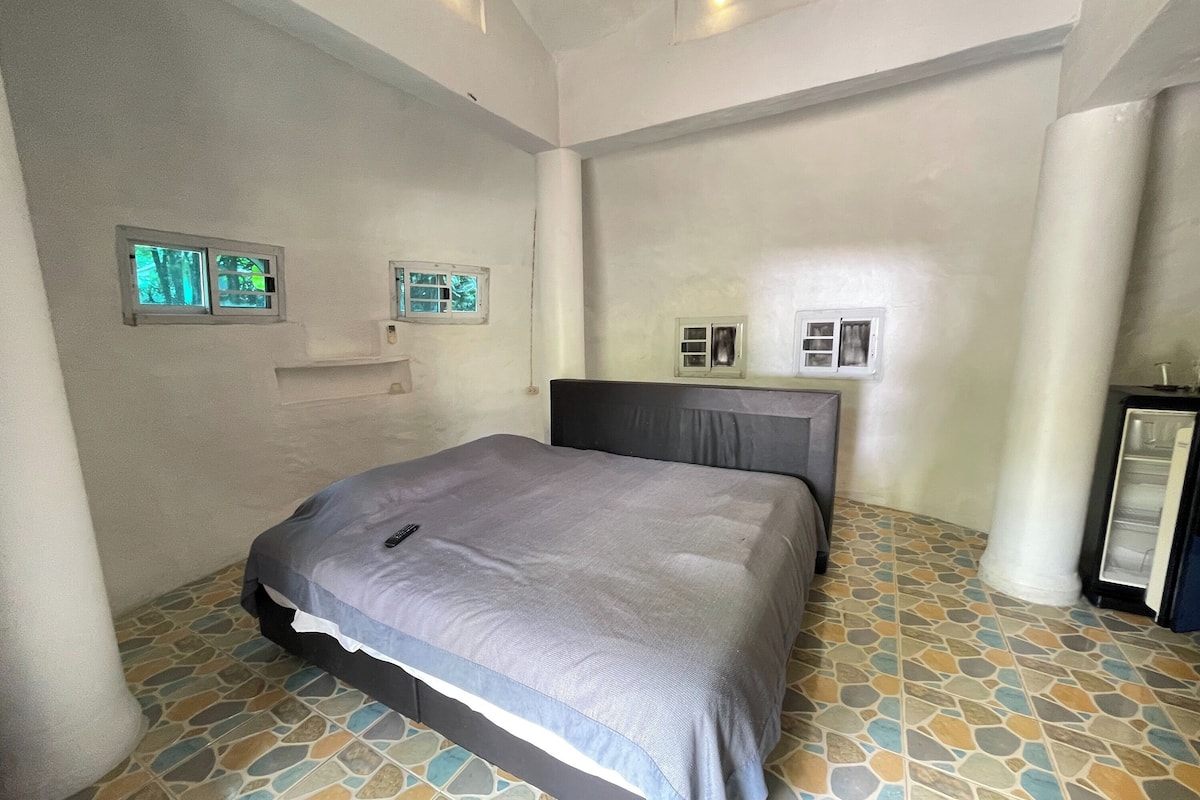 A bedroom with a bed, a small window, a wall-mounted shelf, and a refrigerator.