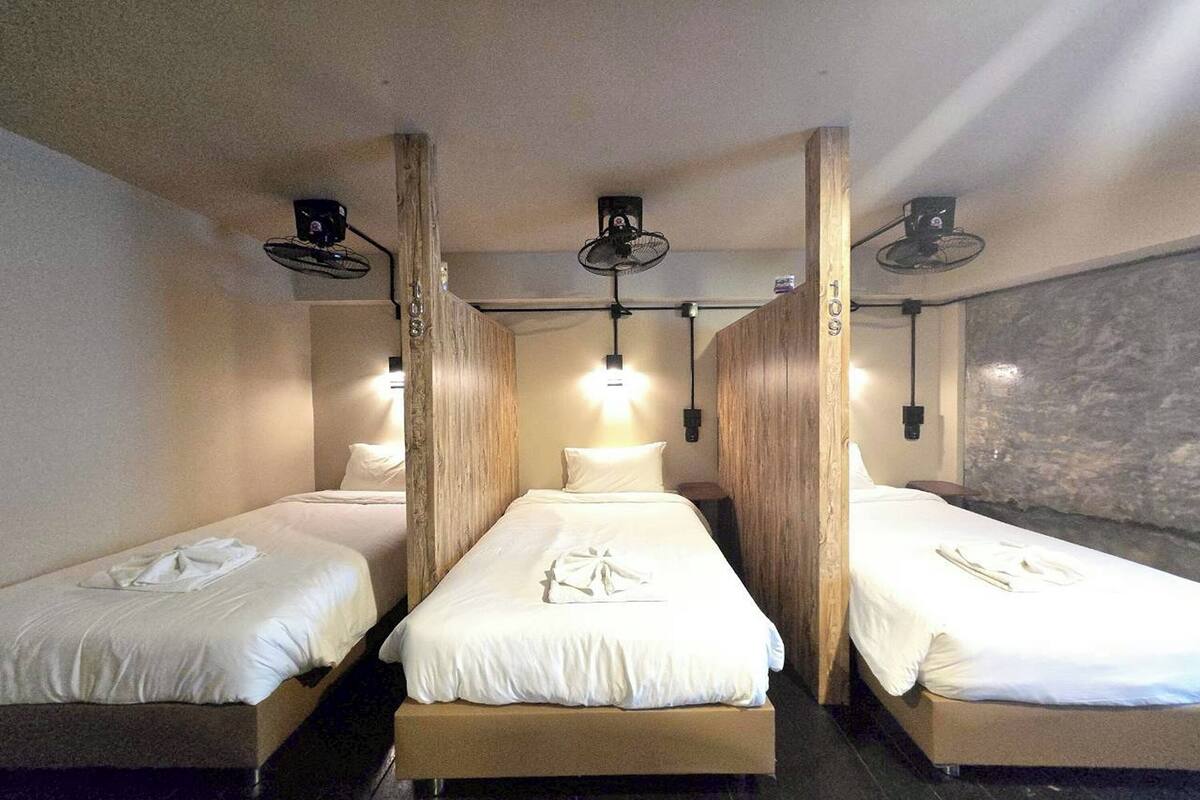 A hotel room with three single beds, each with a white bedspread and a towel. There are ceiling fans and wall-mounted lights.