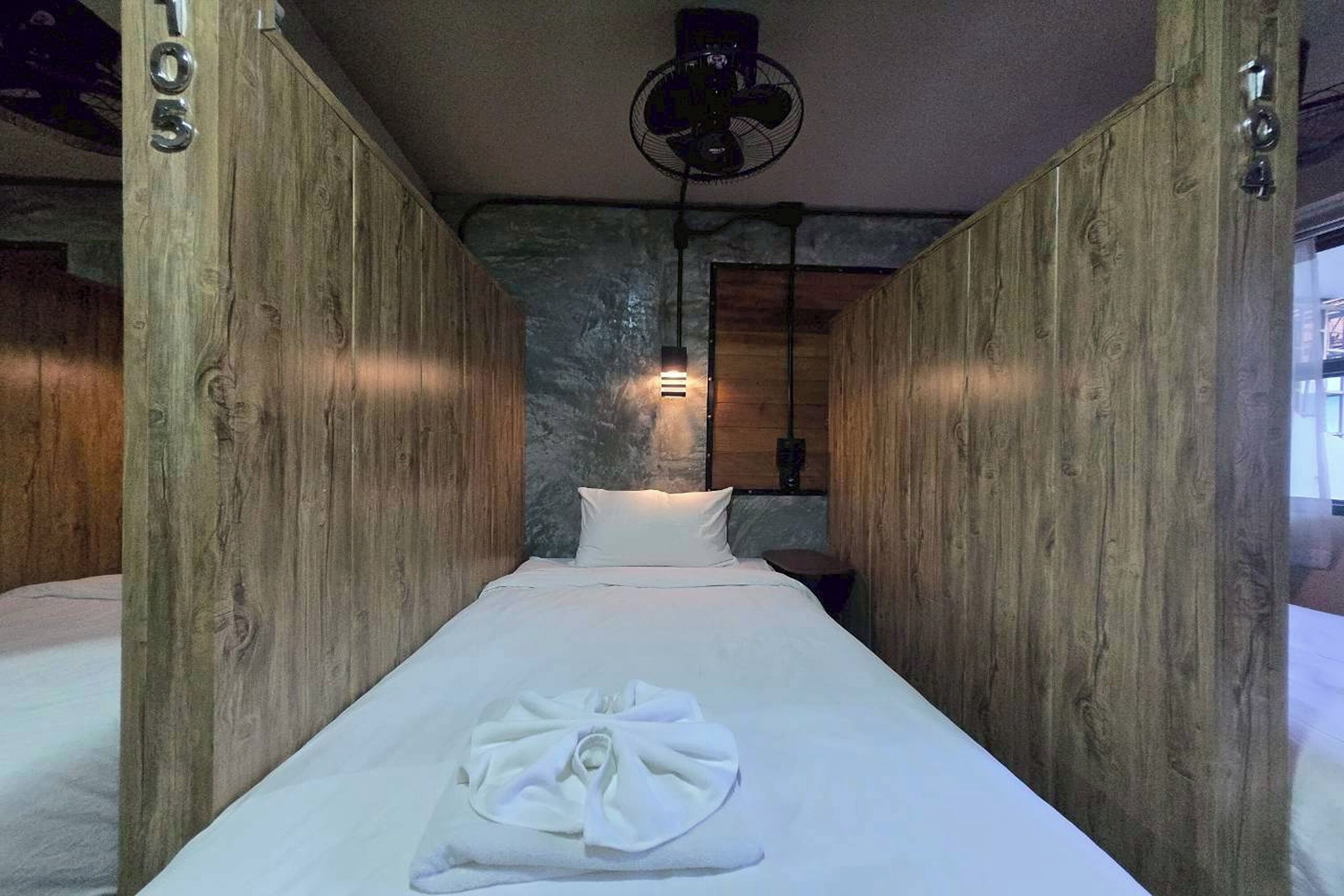 Habitació estreta amb panells de fusta, un llit al final, un ventilador a sobre i una taula petita amb una làmpada.
