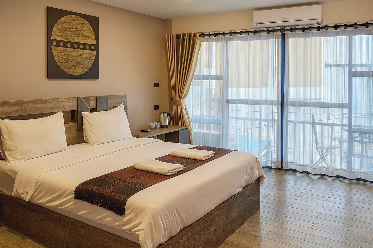 A neatly made bed with white linens and a brown throw blanket, a wooden headboard, and a large window with sheer curtains.