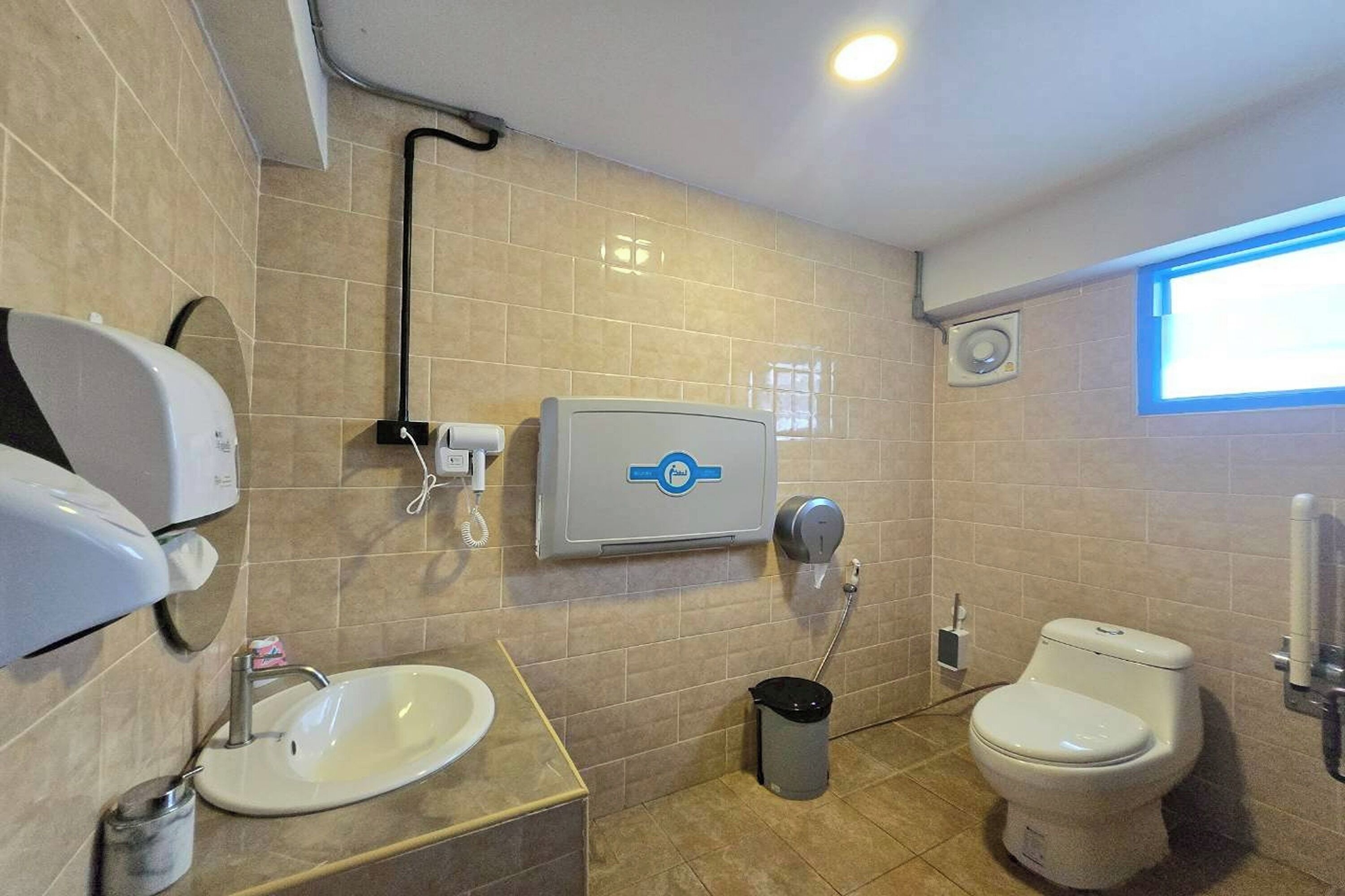 A bathroom with a sink, toilet, hand dryer, and a trash bin.