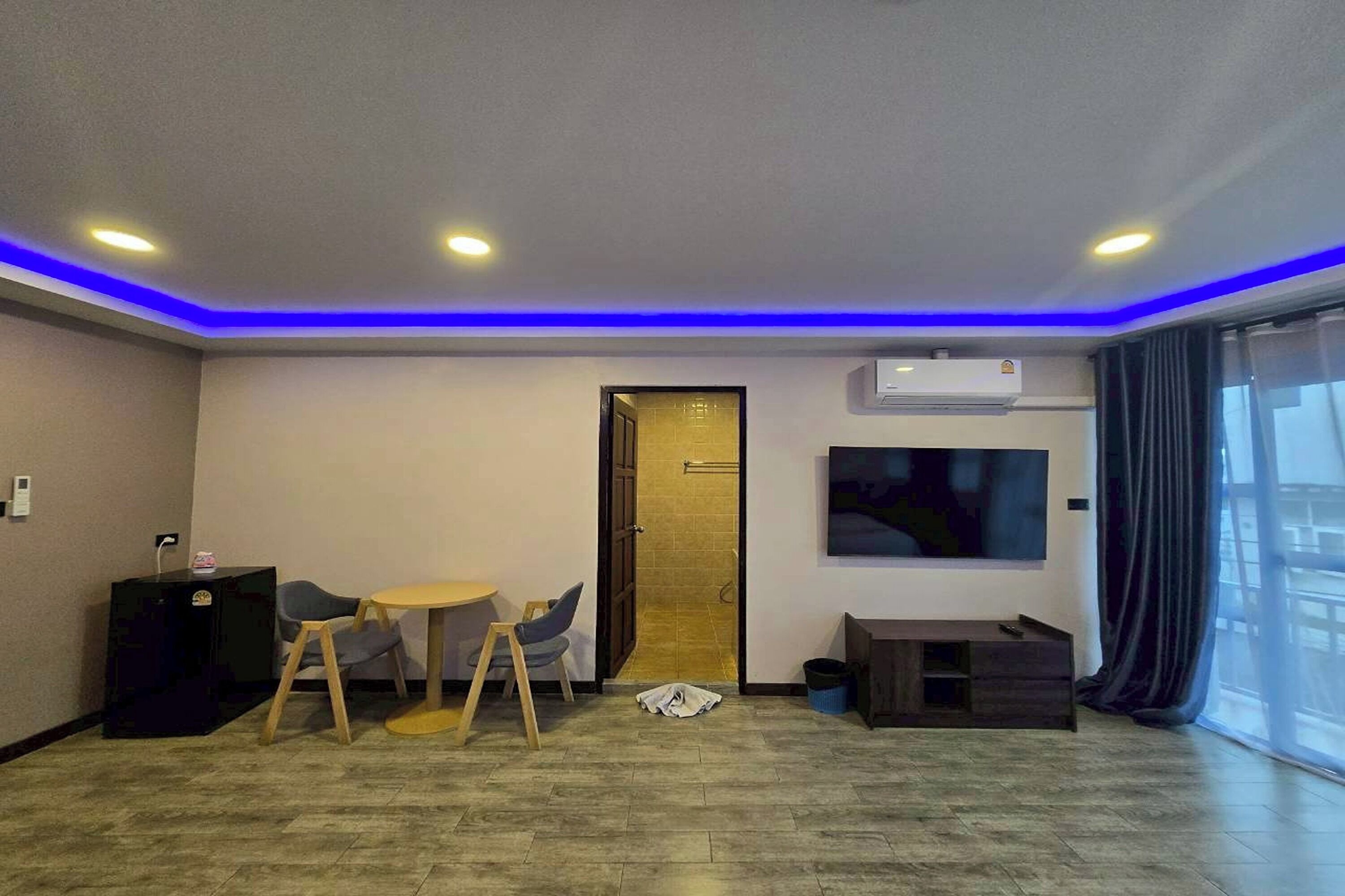 Una habitación de hotel moderna con comedor, televisor montado en la pared y un amplio ventanal con cortinas.