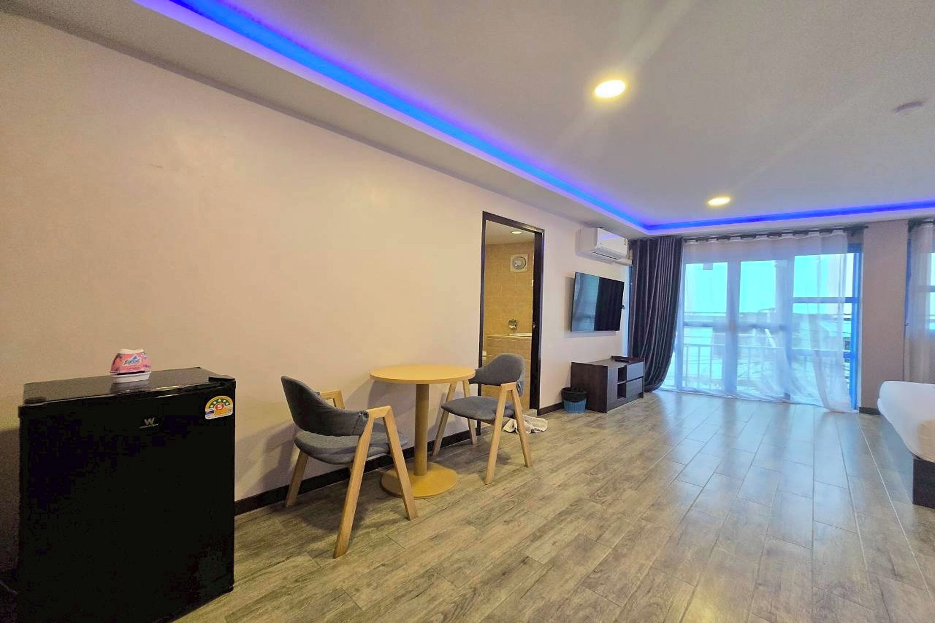 A hotel room with a wooden floor, a small round table with two chairs, a TV on the wall, and a bed with a white mattress.
