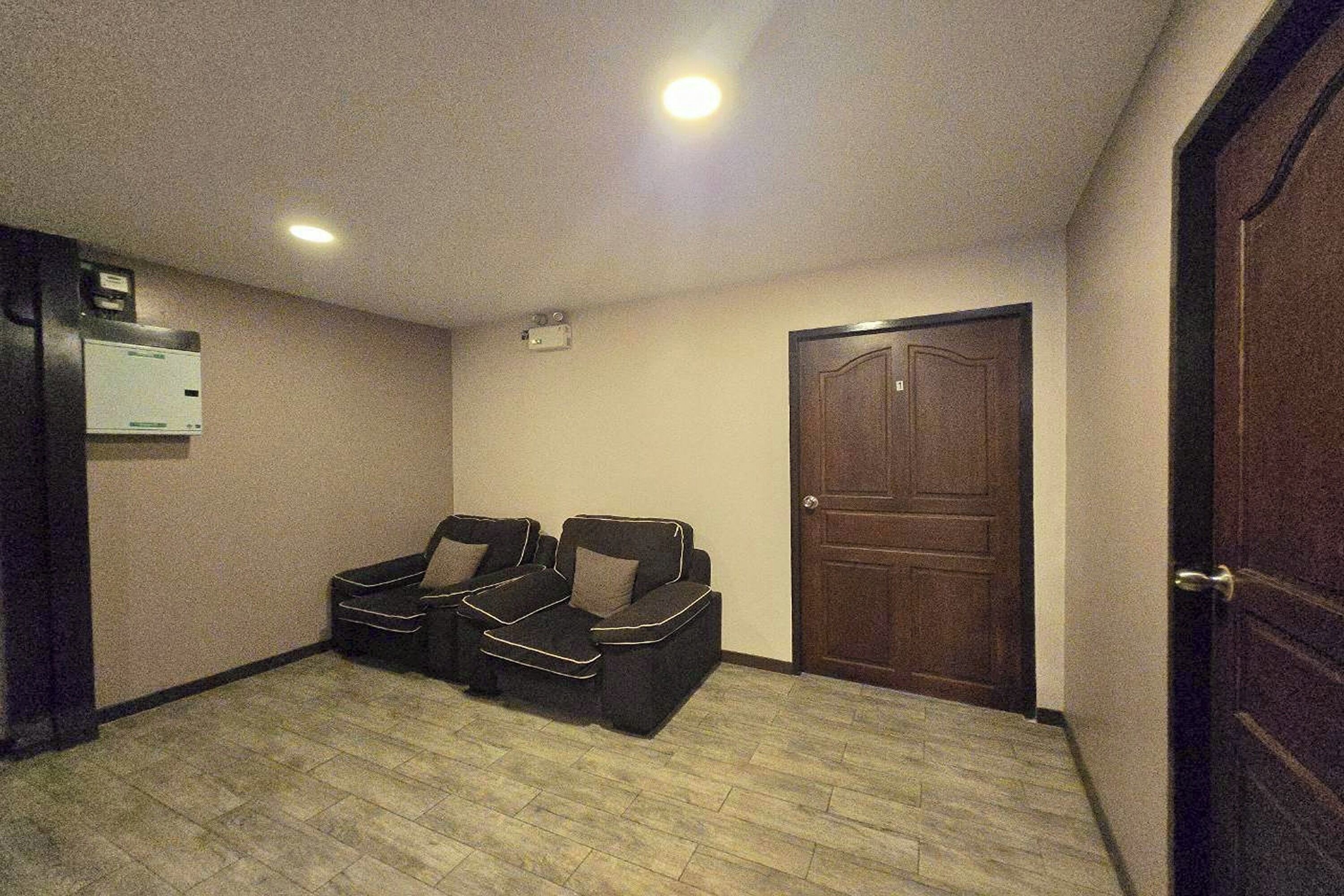 A room with a wooden door, a wall-mounted air conditioning unit, and a pair of black sofas with cushions.