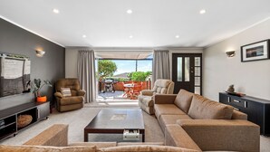 A modern living room with a sofa set, a coffee table, and a TV. There is a balcony with outdoor furniture and a view of greenery.