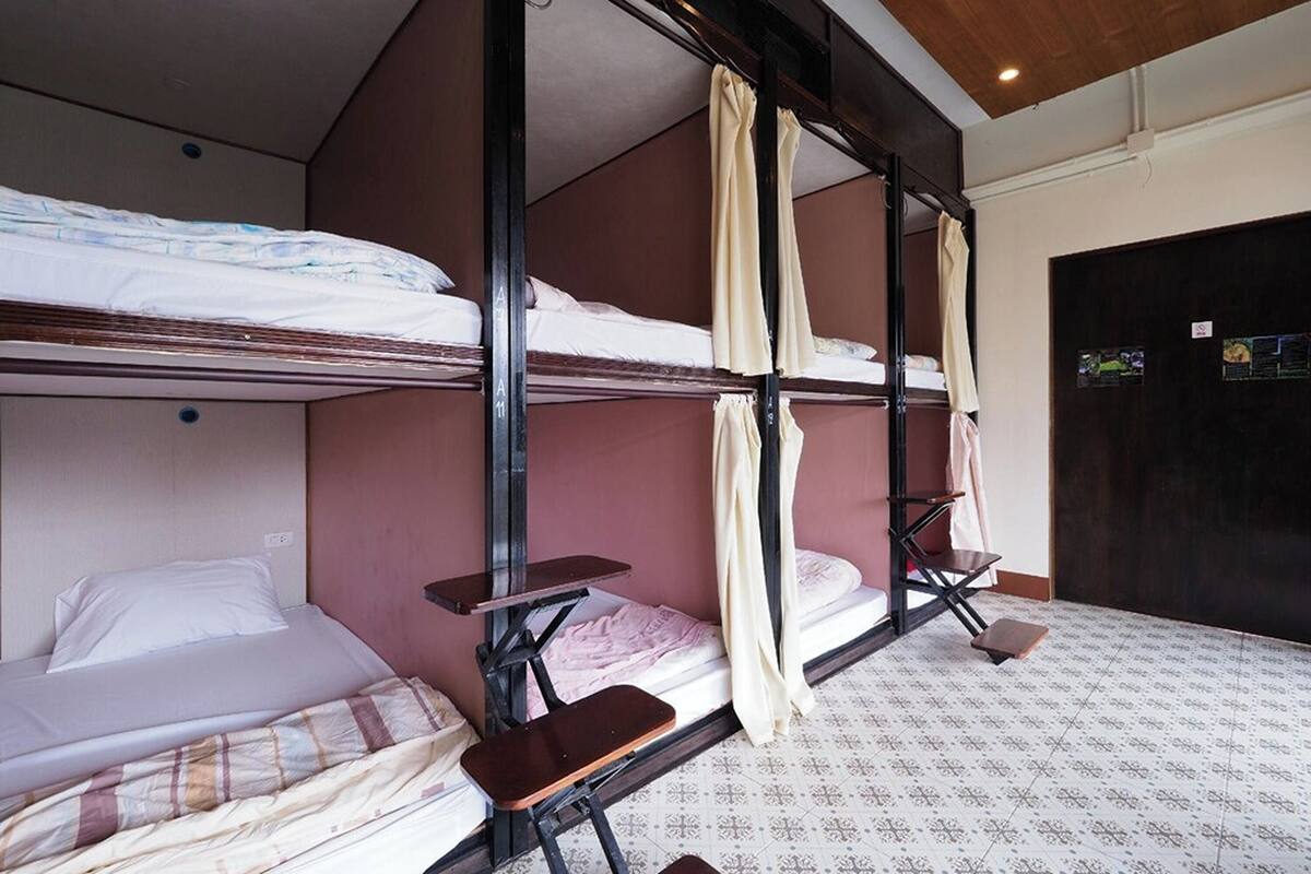 Habitación con literas, mostrando tres camas con estructura de madera y colchones. Se observan bancos de madera adosados a las camas. El suelo tiene baldosas con diseño y una puerta oscura.