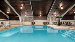 An indoor swimming pool with a wooden ceiling, clear glass windows, and multiple chairs around the pool.