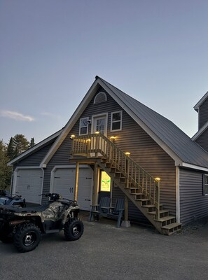 A two-story house with a wooden staircase and a balcony, two garage doors, and two ATVs parked in front.