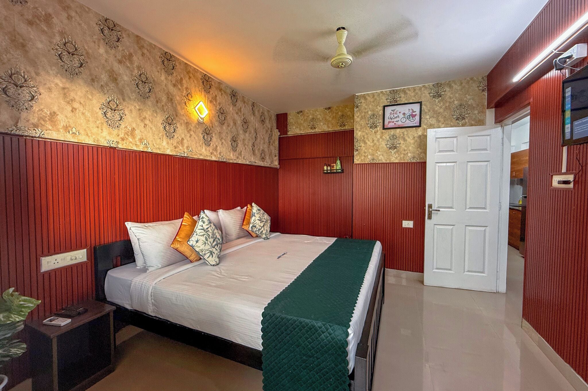 A hotel room with a bed, a nightstand, a green throw blanket, a ceiling fan, and a door leading to another room.