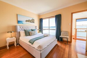 Une chambre d’hôtel avec un grand lit, une table de chevet, une chaise, un tableau et une vue sur la mer par la fenêtre.