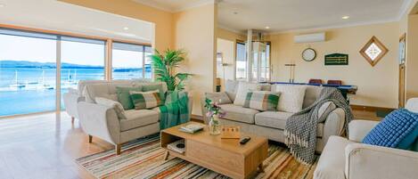 A spacious living room with a large window overlooking a body of water, a sofa set, a coffee table, and a potted plant.