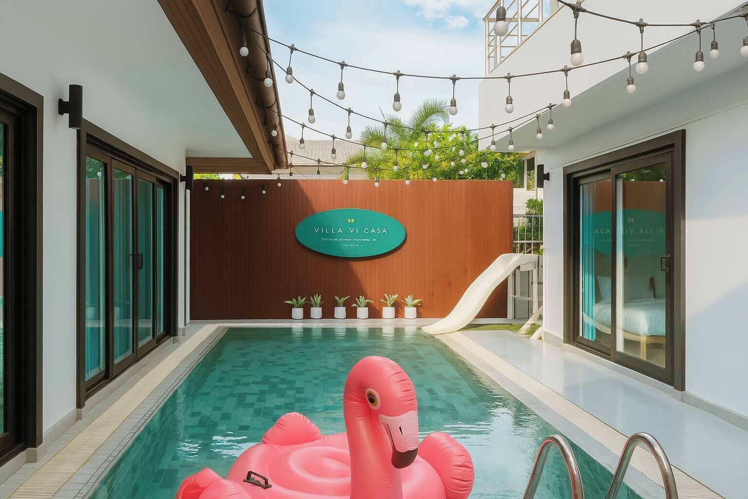 A swimming pool with a slide, a pink inflatable flamingo, and a sign with the name 'Villa Vi Casa'.