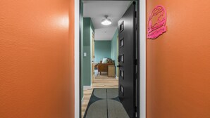A hallway with an orange wall, a black door, and a doormat leading to a room with a bed and a chair. - Cozy 2 Bedroom Apartment within walking distance from the Hospital. (Williston)