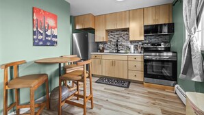 A compact kitchen with wooden cabinets, a refrigerator, a microwave, and an oven. There's a dining area with a table and chairs, and a painting on the wall. - Cozy 2 Bedroom Apartment within walking distance from the Hospital. (Williston)