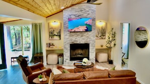 Una sala de estar con chimenea de piedra, un televisor montado encima y un ventanal con vistas a un área boscosa.