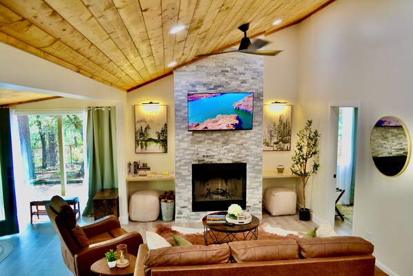 Una sala de estar con chimenea de piedra, un televisor montado encima y un ventanal con vistas a un área boscosa.