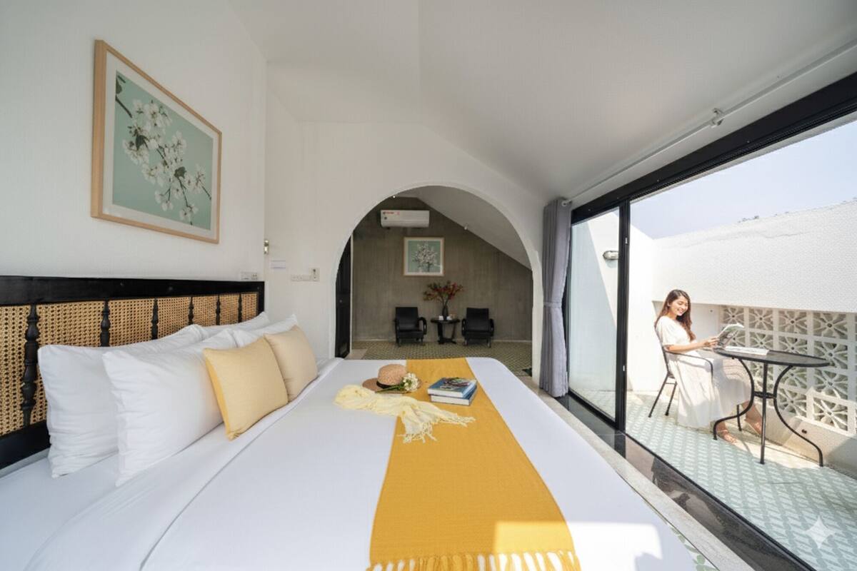 Habitación de hotel con una cama grande, un plaid amarillo, dos sillones, una mesita y un balcón donde hay una persona sentada.