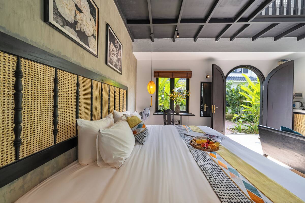 Habitación de hotel con cama, cabecera de ratán, ventilador, ventana con vista a follaje y una bandeja de desayuno sobre la cama.