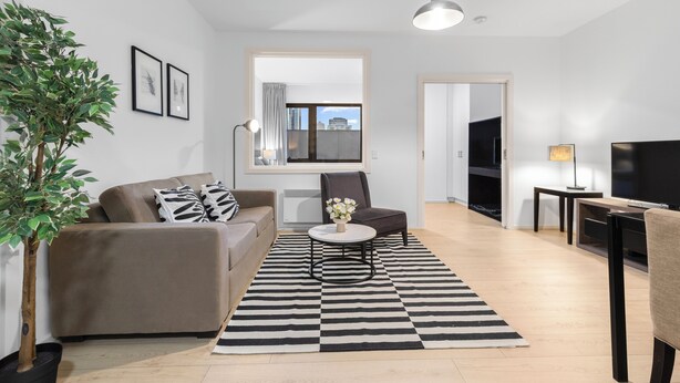 A modern living room with a sofa, armchair, coffee table, and a potted plant. There is a black and white striped rug on the floor. - 
Central Emporium | Hotel-Style 1BR w/ King Bed (Melbourne)