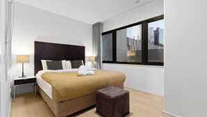 A modern hotel room with a large bed, a small bedside table, a nightstand, a window with a city view, and a wooden floor. - 
Central Emporium | Hotel-Style 1BR w/ King Bed (Melbourne)