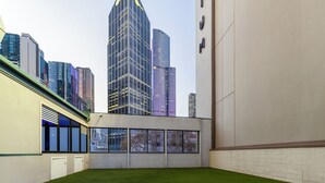 A room with a green artificial turf floor, large windows, and a view of tall buildings in the background. - 
Central Emporium | Hotel-Style 1BR w/ King Bed (Melbourne)