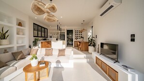 A modern living room with a sofa, coffee table, and TV. There are shelves with decorative items and a kitchen in the background. - Villa Mulia Uluwatu (Pecatu)