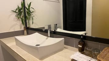 Un baño con un lavamanos blanco, un espejo, un perchero para toallas, una planta en maceta y una botella de jabón para manos.