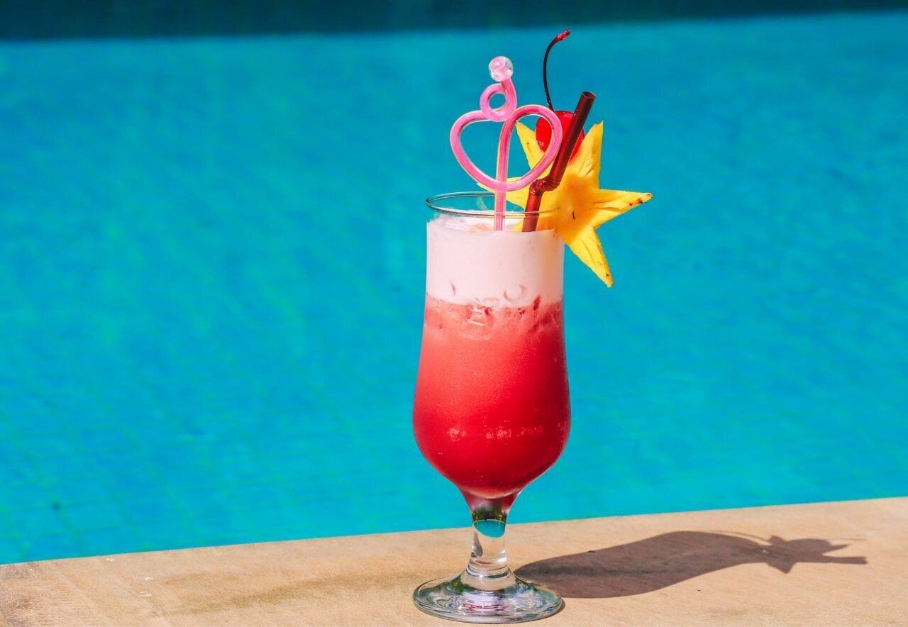A cocktail in a tall glass with a pink heart-shaped topper and a yellow star decoration, placed on a wooden surface with a pool in the background.