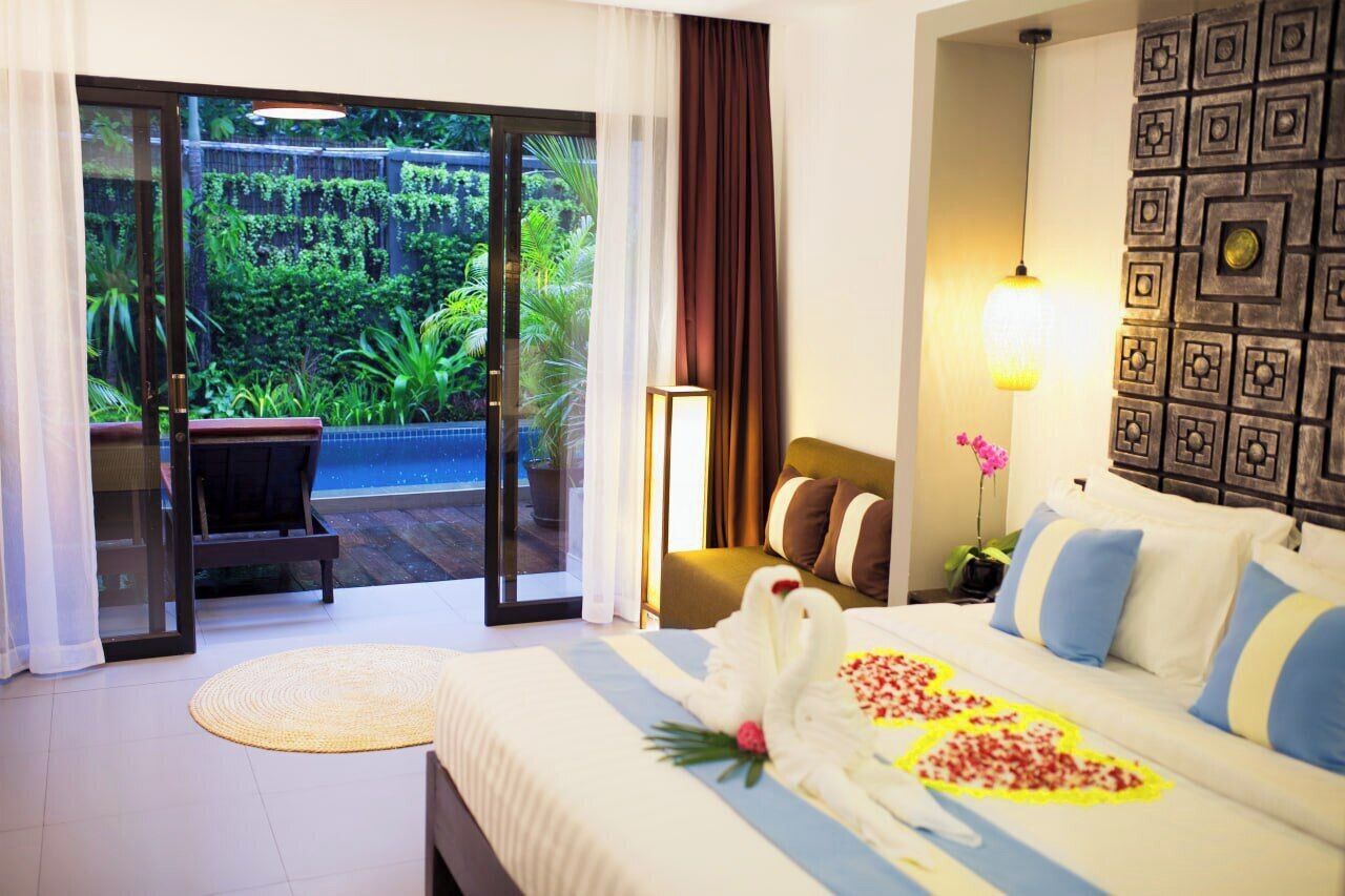 A hotel room with a bed, a chair, a view of a lush garden through a sliding glass door, and a wall with a geometric pattern.