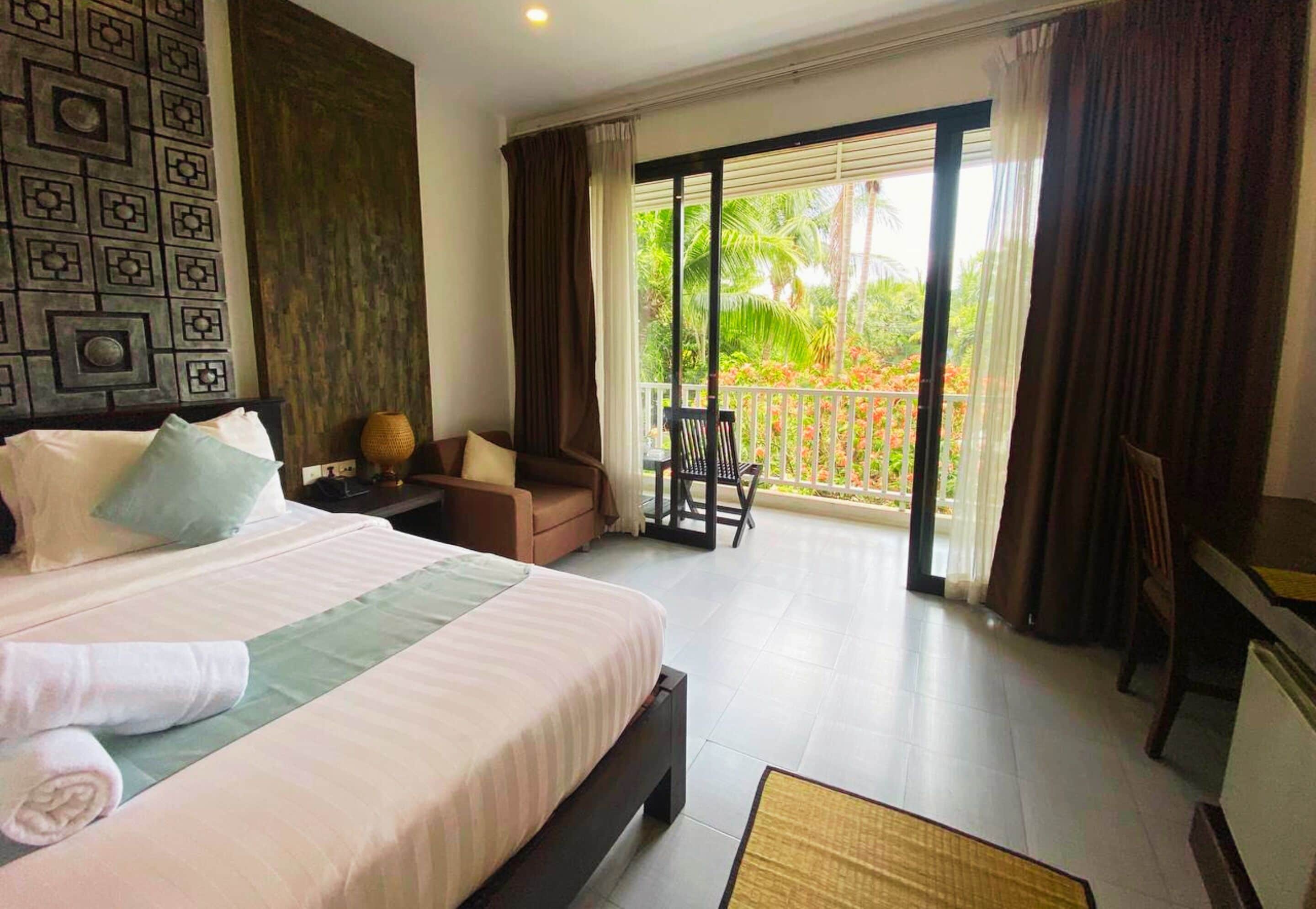 A hotel room with a large bed, a desk, a chair, and a balcony with a view of greenery.