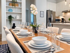 A dining area with a long wooden table set for a meal, white chairs, and a kitchen in the background.