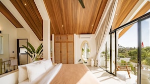 Un dormitorio con una cama grande, techo de madera, ventanas amplias y vistas a la vegetación.