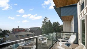 Un balcón con barandas de vidrio, sillas Adirondack blancas y vistas a casas y árboles vecinos.