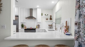 A modern kitchen with white cabinetry, a stainless steel range hood, and a white countertop. There are three wicker stools under the counter.