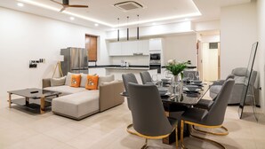 A modern living room with a sofa, dining table, and chairs, a kitchen in the background, and ceiling fans. - 3Bed SeaViewVilla FishermanVillage KBR43 (Koh Samui)