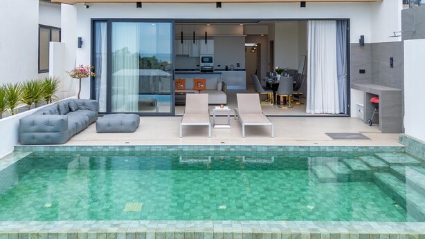 A modern outdoor pool area with lounge chairs, a sofa, and a kitchen visible through glass doors. - 3Bed SeaViewVilla FishermanVillage KBR43 (Koh Samui)