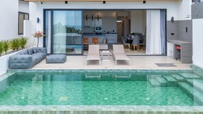 Een modern zwembadgedeelte buiten, met ligbedden, een loungebank en een keuken zichtbaar door glazen deuren.