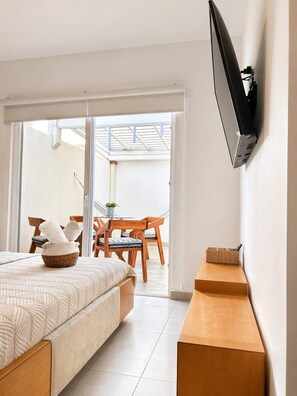 Quarto de hotel moderno com cama, televisão presa à parede, mesa de cabeceira de madeira e varanda com mesa e cadeiras.