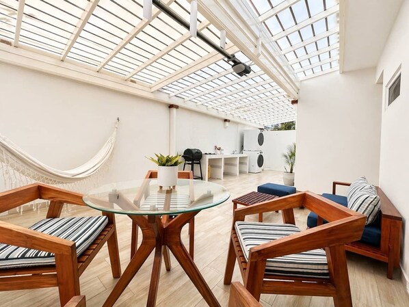 Uma sala de estar integrada com uma mesa de vidro, cadeiras de madeira, uma rede e uma máquina de lavar roupa.