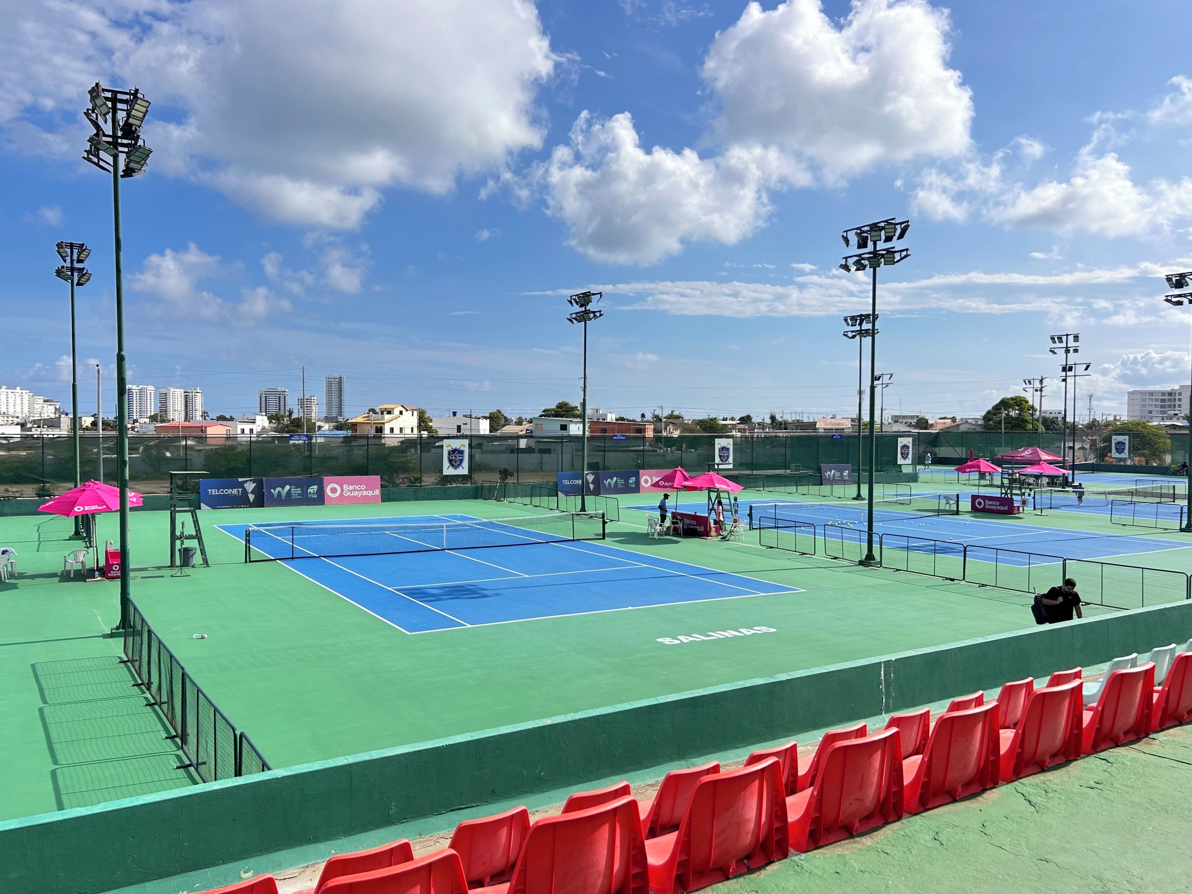 A tennis court with multiple courts, seating areas, and floodlights.